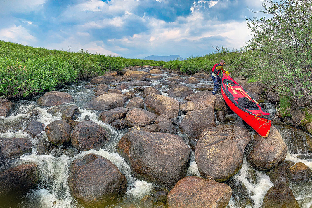man portages a canoe along a bouldery stream bed at the start of his Arctic expedition