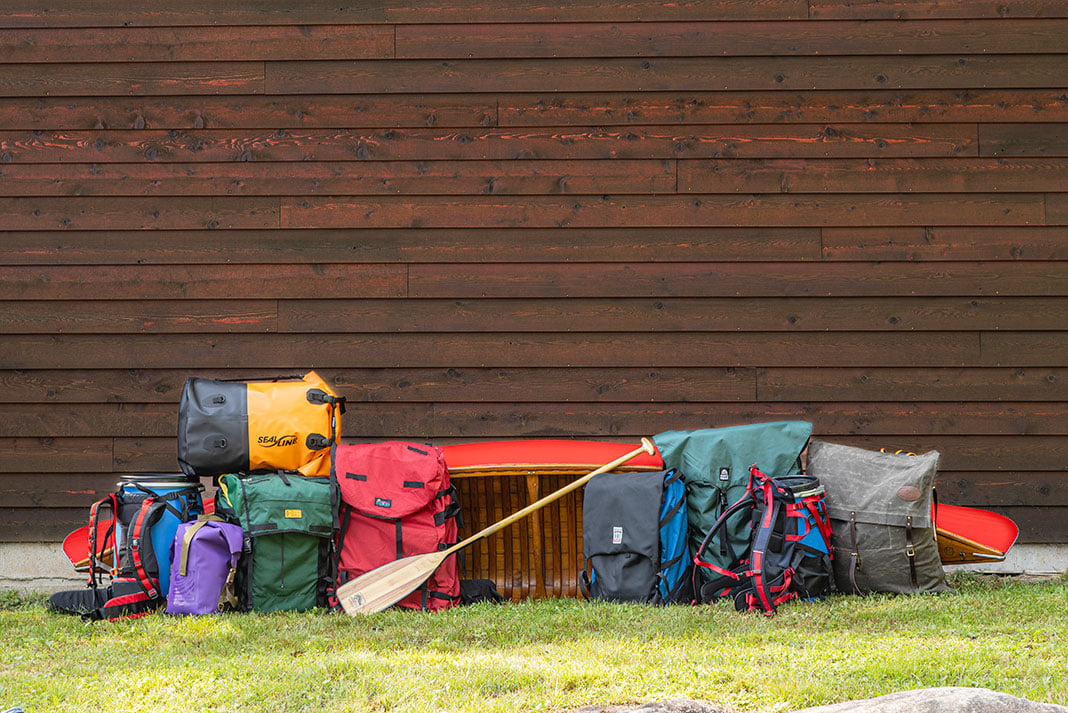 a selection of canoe packs and barrel harnesses in front of a red wood-canvas canoe and wood slat wall
