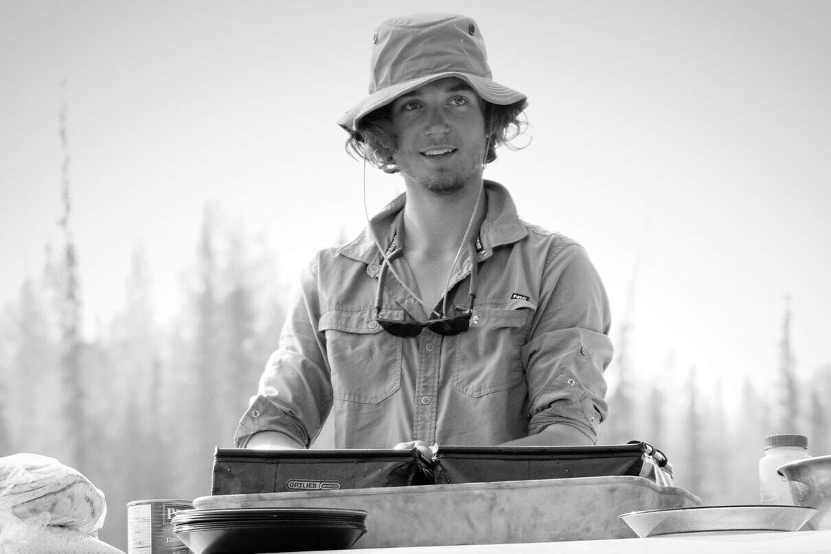 black & white photo of a young man in bucket hat washing dishes at a camp kitchen set up on an overturned canoe