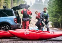 three whitewater kayakers gear up by the trunk of their vehicle with red kayaks on the ground in front