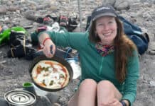 woman sitting near campfire holds up a cast iron skillet with backcountry pizza baked inside