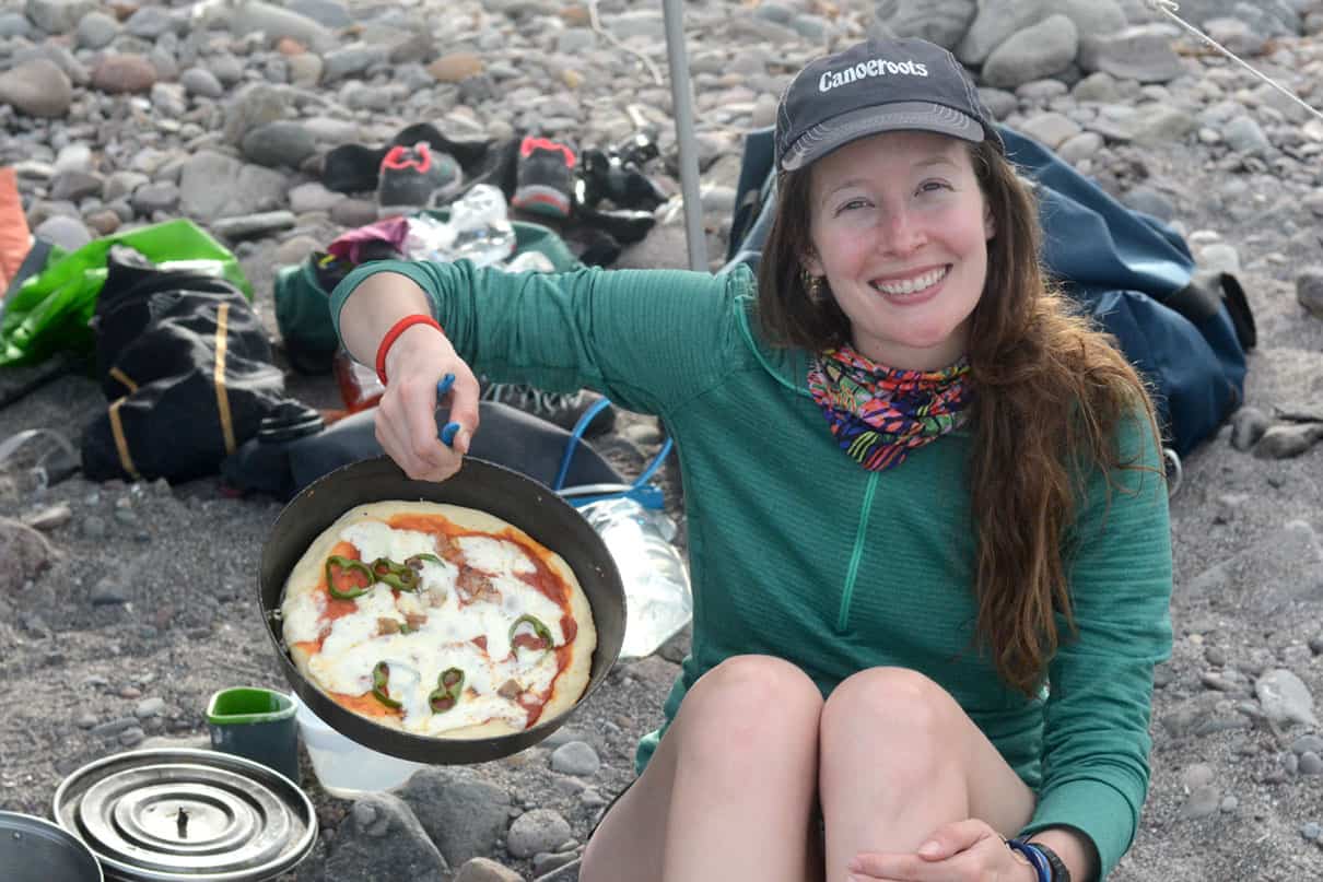woman sitting near campfire holds up a cast iron skillet with backcountry pizza baked inside