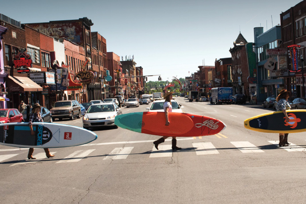 Three people crossing road with paddleboards under their arm.