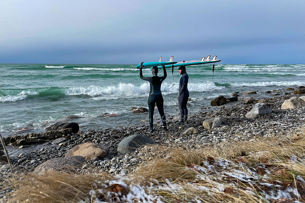 a man and woman in wetsuits stand on a rocky, wave-strewn shore balancing surboards on their heads