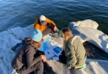 three women gather around a paper map spread out on rocks near the water, rather than using navigation apps