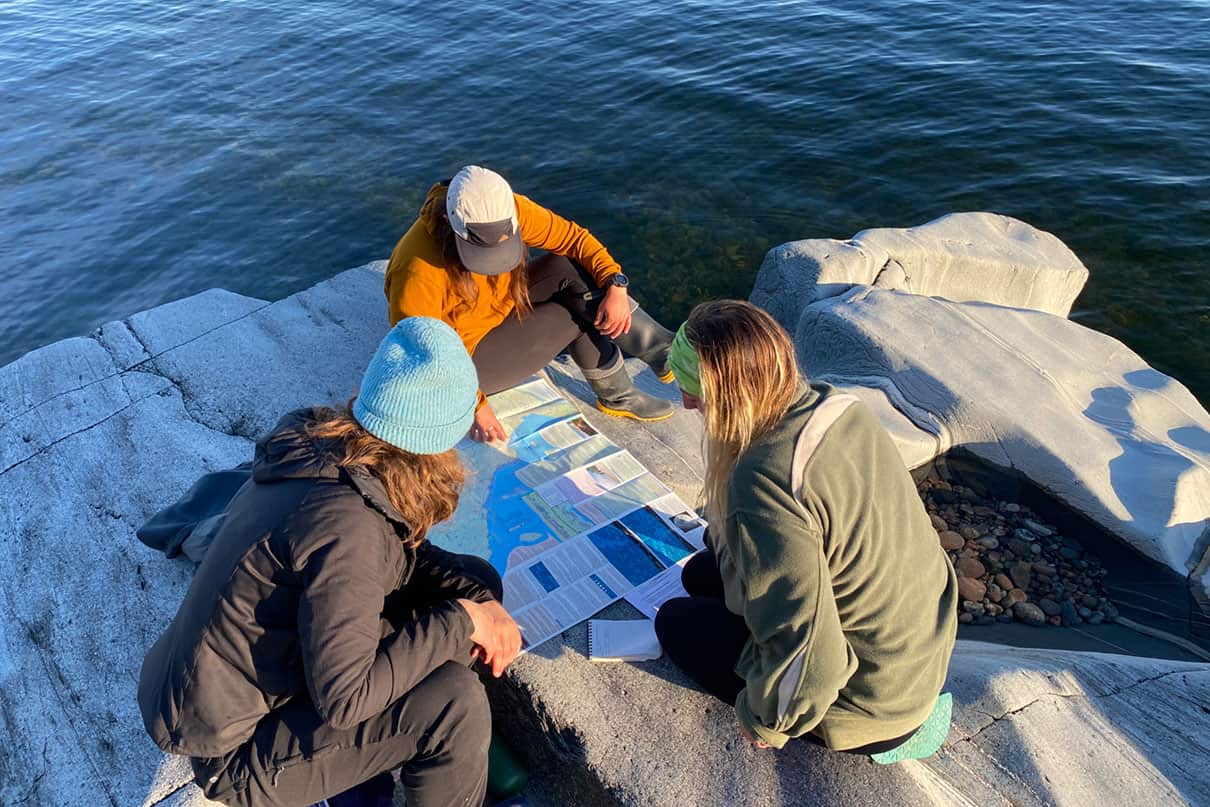 three women gather around a paper map spread out on rocks near the water, rather than using navigation apps