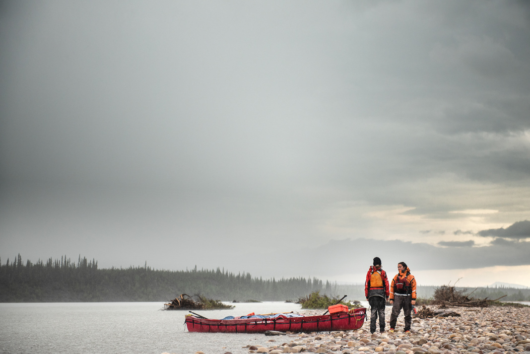 Two canoeists stand at the side of a lake against a grey sky