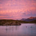 Two touring kayakers paddle in pink dawn or dusk light with mountains behind them